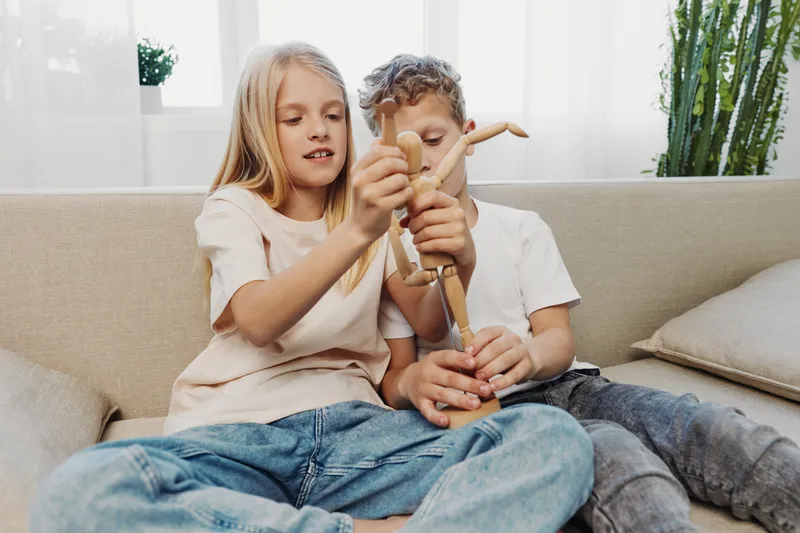 Two children sitting on a couch playfully adjusting a wooden artist mannequin in a bright modern living room with natural light and green plants in the background.
