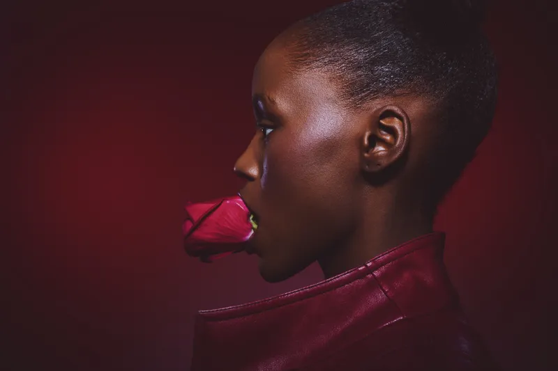 Profile of a dark skinned African woman holding a red rose in her mouth wearing a red leather jacket. Concept of beauty, fashion, and elegance on red background.