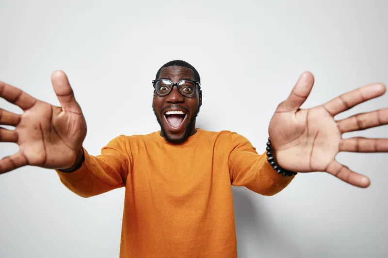 African man with dark skin and glasses wearing orange sweater smiling and reaching out with open hands for hug or welcome on white background.