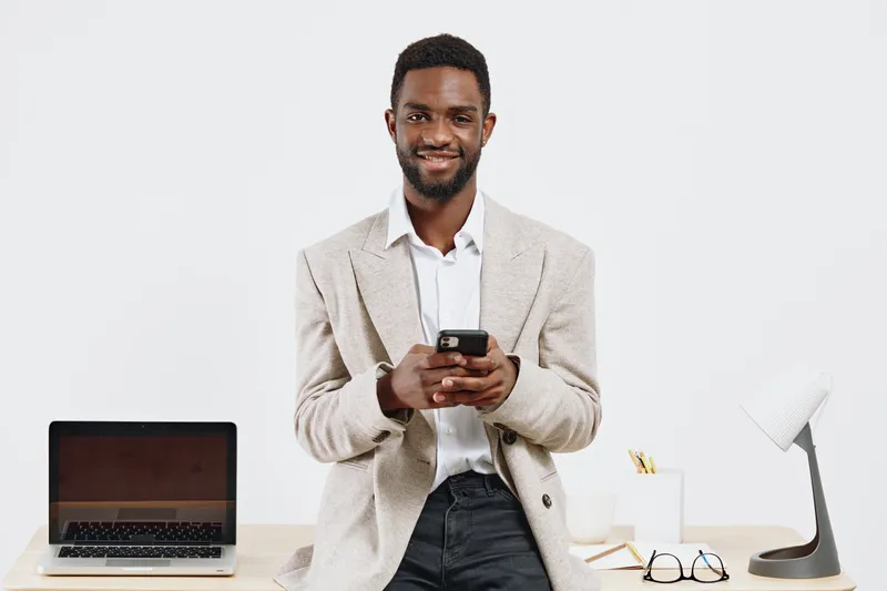 Confident smiling man in casual beige blazer and white shirt using mobile phone in bright office setting with desk, laptop, and desk lamp. People lifestyle concept with modern workspace ambiance.