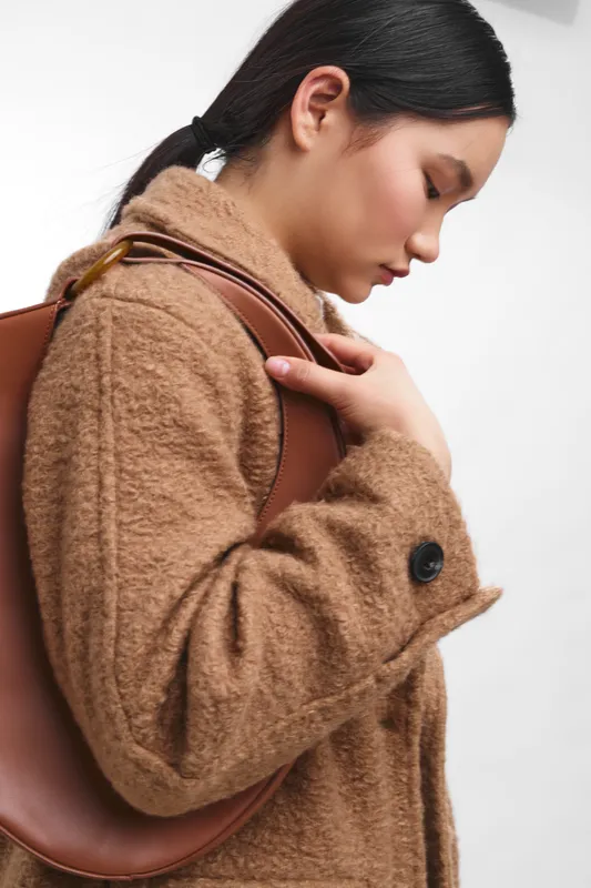 Asian woman in a warm coat stands with a brown shoulder bag, gazing downward with a thoughtful expression in a clean studio, radiating calm, sophistication and quiet focus.