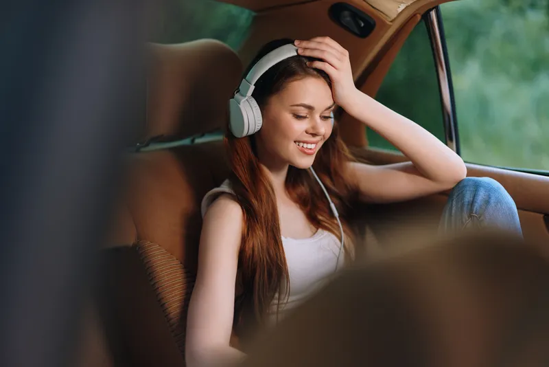 Young woman with headphones enjoying music in a car, smiling and relaxed, featuring a natural background and warm color palette.