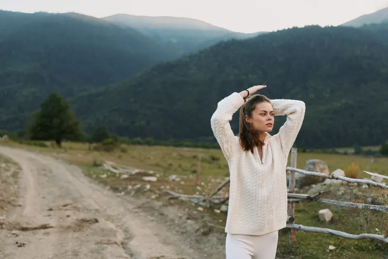 A woman in a cream sweater stands outdoors among rolling mountains, hands in hair, conveying a calm, reflective moment in nature and a sense of freedom.