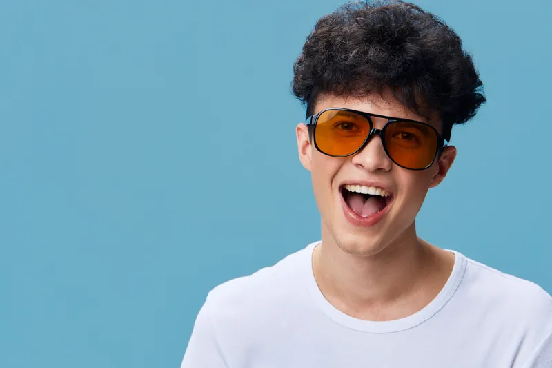 smiling man with curly hair wearing orange sunglasses and white t-shirt laughing joyfully against blue background, youthful happy expression, casual style