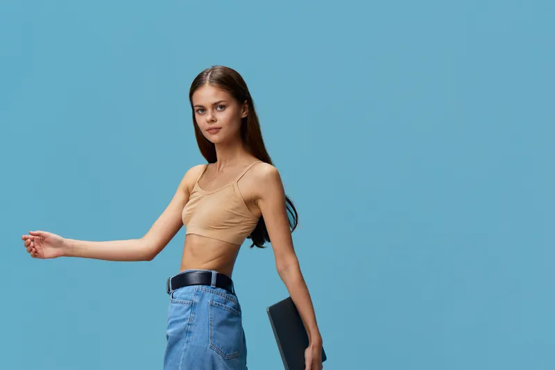 young woman holding laptop in casual outfit with beige crop top and denim skirt against blue background, modern and stylish tech lifestyle concept