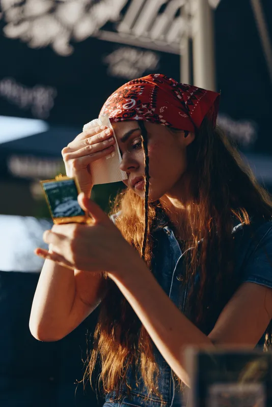 Lifestyle woman wearing modern boho-western denim street style outfit, applying makeup outdoors in warm film color tones with natural sunlight and relaxed mood.