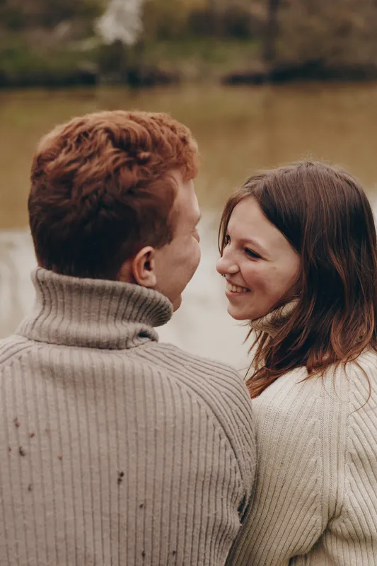 Joyful couple outdoors, wearing cozy sweaters, share a warm smile by a tranquil lake, embracing in soft light, natural colors, gentle breeze, intimate connection, candid moment