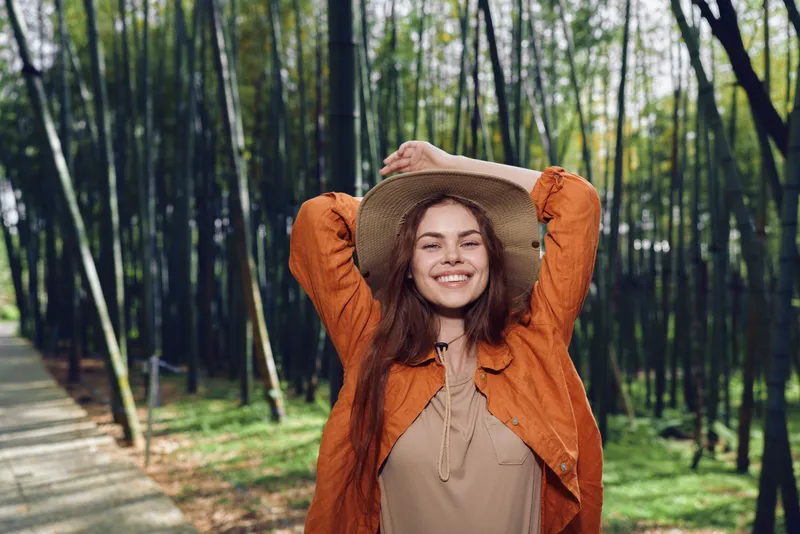 Woman smile hat forest nature portrait happy outdoors: young woman in a straw hat and orange jacket stands on a tree-lined path, raising her arms and smiling in warm sunlight.