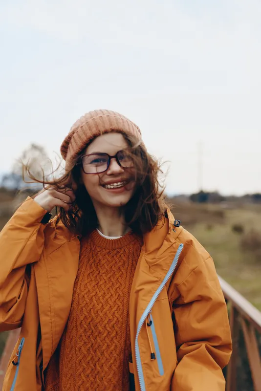 Woman lifestyle outdoors, smiling in hat and glasses, wearing mustard jacket and knit sweater by road railing in autumn countryside. Warm daylight, relaxed travel mood.