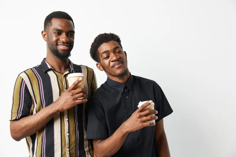 Two black men enjoying coffee together, smiling brightly in casual clothing. Capturing friendship and joy in a relaxed setting, ideal for lifestyle themes.