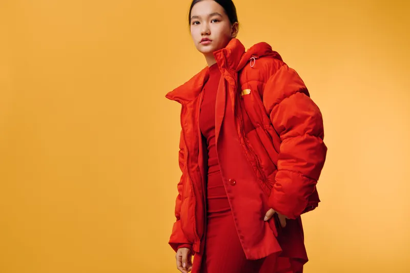 Young woman wearing red puffer jacket and matching outfit posing against orange background. Fashion model in warm clothing with confident expression and modern style portrait.
