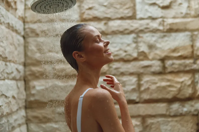 woman showering outdoors under water droplets with wet hair and closed eyes, enjoying refreshing bathing routine near beige stone wall