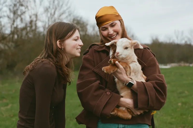 Two women outdoors share a warm moment with a gentle goat kid, affection and laughter on a rural field, casual jackets, natural light, candid lifestyle scene, friendship and animal bond.