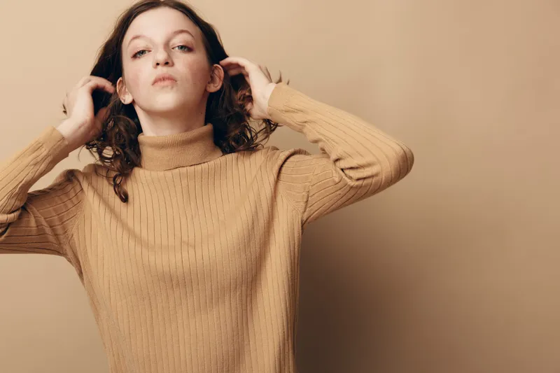 Young woman with curly hair wearing beige knitted sweater poses confidently on beige background. Concept of fashion, casual style, autumn clothing, and natural beauty portrait.