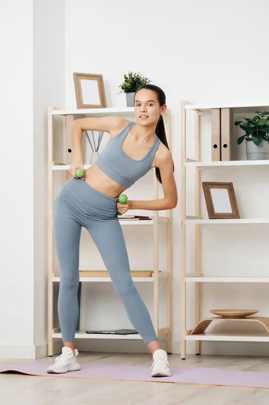 Fitness woman in blue athletic wear performs a side bend with dumbbells at home, a neat studio with shelves, plants, and framed decor; a focused workout and cardio.