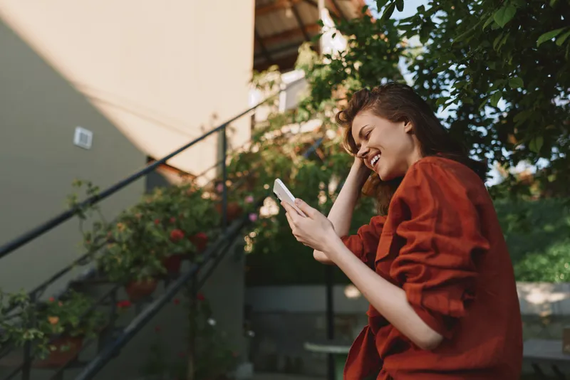 A woman outdoors smiles at her smartphone, wearing a red sweater. A garden balcony scene with greenery conveys casual lifestyle and everyday joy