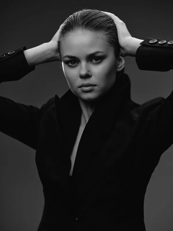 Black and white portrait of a serious woman with slicked-back hair wearing a black jacket posing on dark background