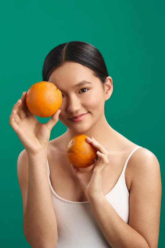 Asian woman holds two oranges against a green backdrop, smiling gently as she presents fruit with a playful pose, creating a fresh, vibrant mood and a healthy lifestyle vibe.
