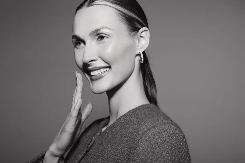 Smiling caucasian woman with light skin and long hair touches her face gently, wearing earrings and textured sweater. Studio portrait with soft lighting and positive mood.