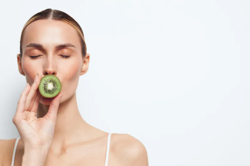 Young woman with closed eyes holding a fresh kiwi slice near her lips, natural skin, minimal makeup, white background, healthy lifestyle, fresh fruit concept