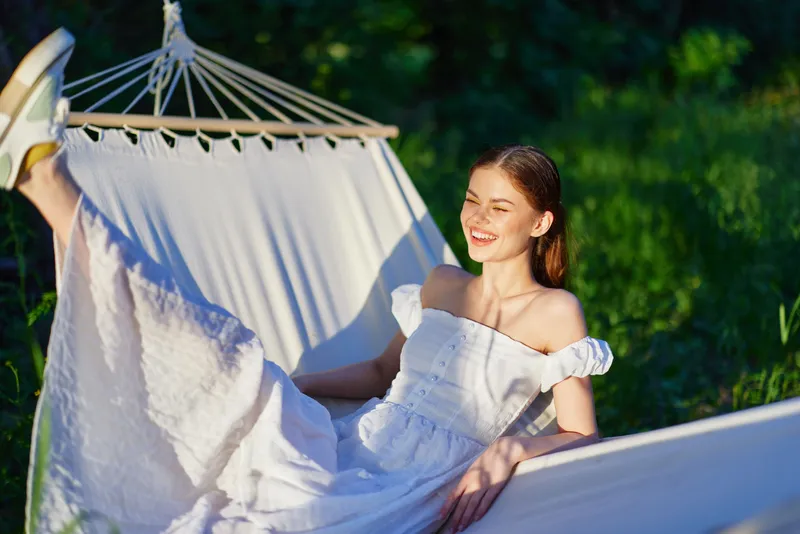Young woman in a white dress relaxing in a hammock, smiling in a lush green garden, evoking a sense of tranquility and summer joy, perfect for wellness themes