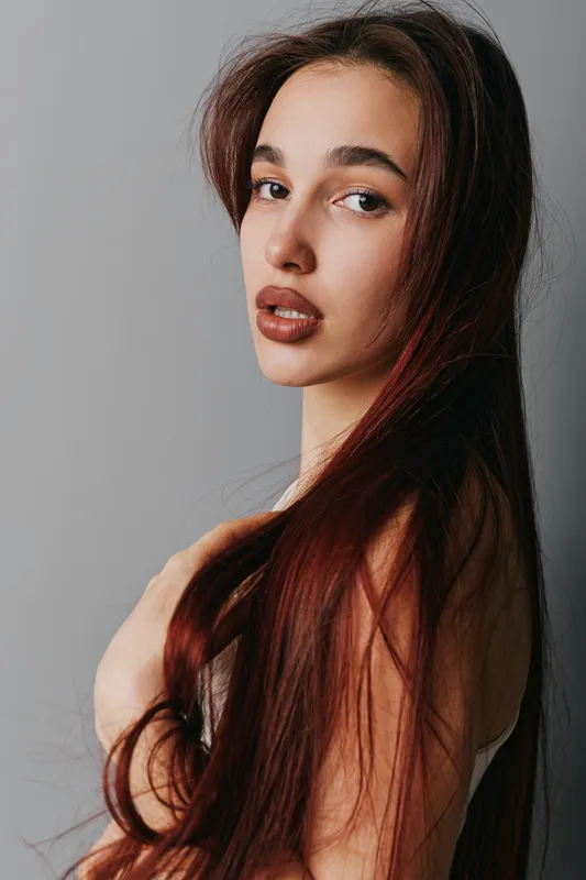 Portrait of young woman with long brown hair looking at camera against gray background. Beauty model with natural makeup and smooth skin in studio lighting for fashion or lifestyle.