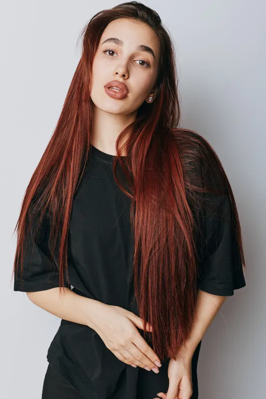 Woman with long red hair and black shirt posing against white background, looking at camera with neutral expression. Portrait of young female model with natural makeup and casual style.