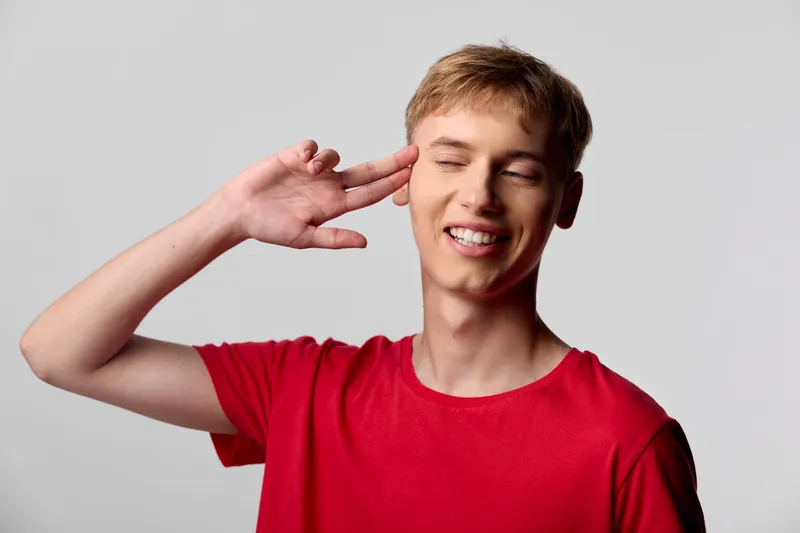 Smiling man in a red shirt with a hand near the ear, suggesting listening and communication in a casual studio setting with soft light and friendly energy