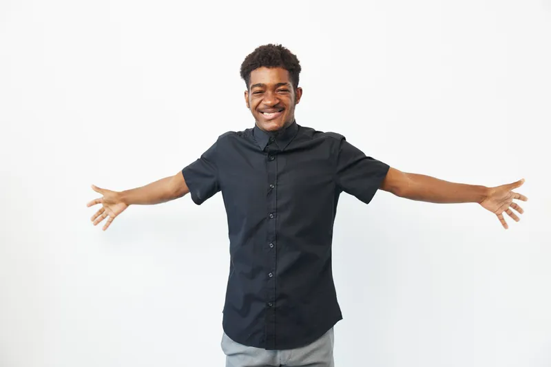 A joyful African American man with arms open wide, smiling brightly against a clean, white background, radiating positivity and enthusiasm in a casual outfit.