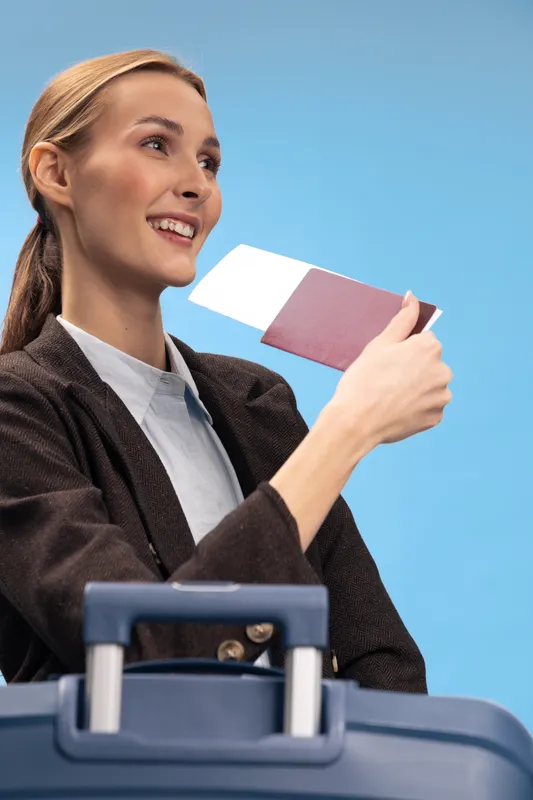 Smiling woman holds boarding pass and passport near suitcase against blue background. Concept for travel, vacation, tourism, and holiday planning with positive mood and destination excitement.