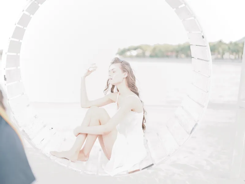 Softly lit portrait of a woman in a white dress sitting inside a circular white prop on a sunlit beach, creating a dreamy, ethereal vibe.