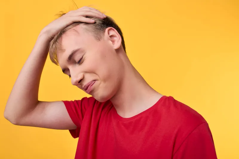 Young man in a red shirt holds his head in frustration against a bright yellow background, conveying stress, fatigue, and discomfort in a candid moment of emotion