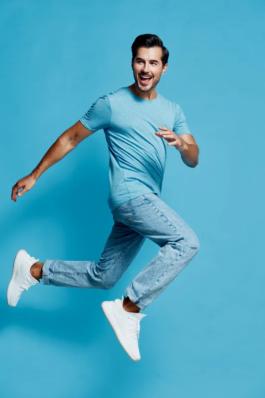 Cheerful young man in blue t-shirt and jeans jumps joyfully while looking at the camera, isolated on vibrant solid blue background. People lifestyle concept