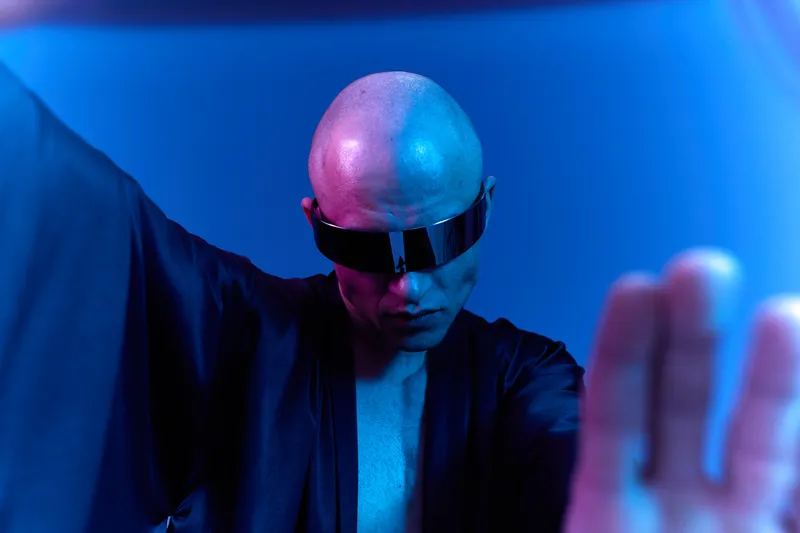 Stylish man in futuristic sunglasses poses dramatically under cool lighting. The image features striking contrasts and a bold, modern vibe that captivates attention.