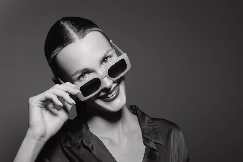 Smiling white woman with fair skin wearing sunglasses and a dark blouse poses against plain background. Concept of fashion, style, confidence, and portrait in monochrome.