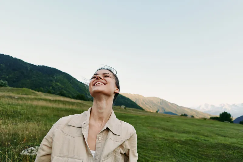 Young woman in a beige jacket stands in a sunlit meadow, looking up with a joyful smile as distant mountains rise, tall grass sways in a light breeze.