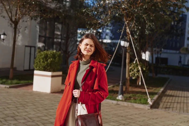 A stylish young woman in a red coat stands outdoors on a sunny day, smiling softly as she poses for a candid street portrait.