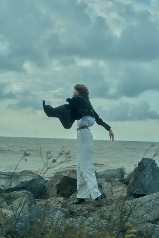 Wind swept figure on rocky coast, flowing coat and white pants, ocean horizon in the background, outdoor fashion and freedom in motion, dramatic sea breeze and bold stance.