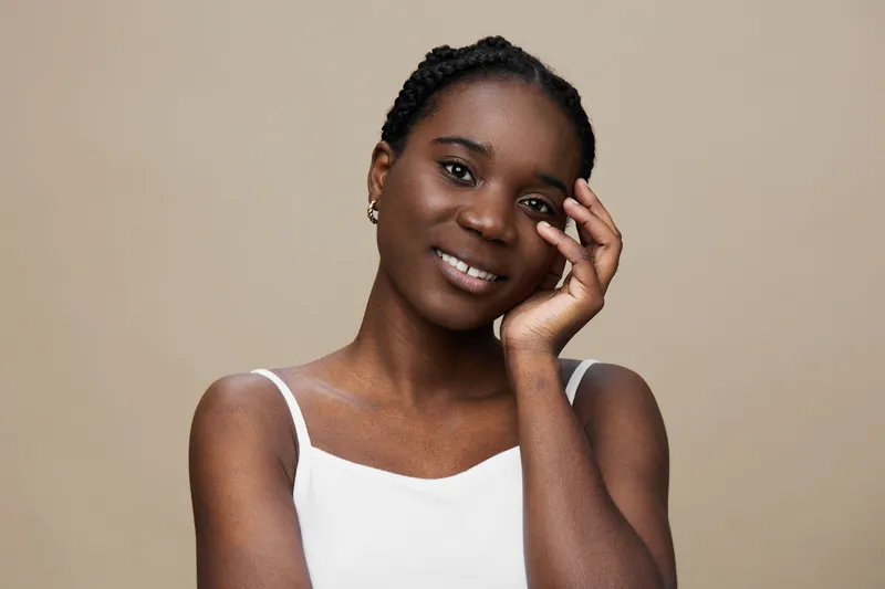 Smiling Black woman in a white top touching her face against a soft beige background, radiating confidence and warmth, perfect for beauty and wellness themes