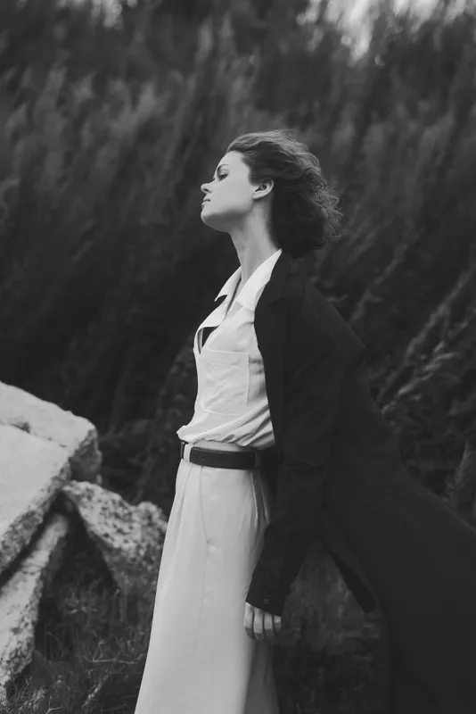 Fashion model outdoors with windblown coat and dress in a monochrome, dramatic editorial look, moody atmosphere and flowing fabrics, elegant stance amid rocky scenery for a timeless image