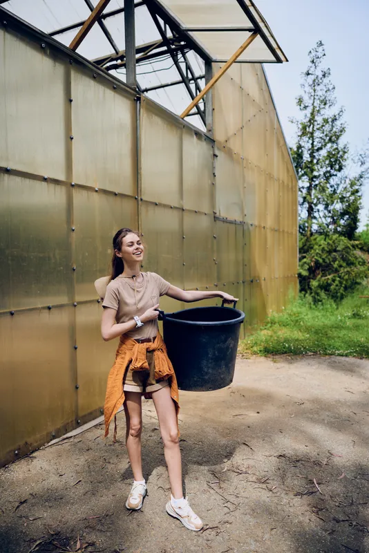Woman holding bucket near greenhouse, smiling in outdoor setting while gardening. Young gardener in casual outfit with tied sweater and sneakers, carrying large plastic container on sunny day.
