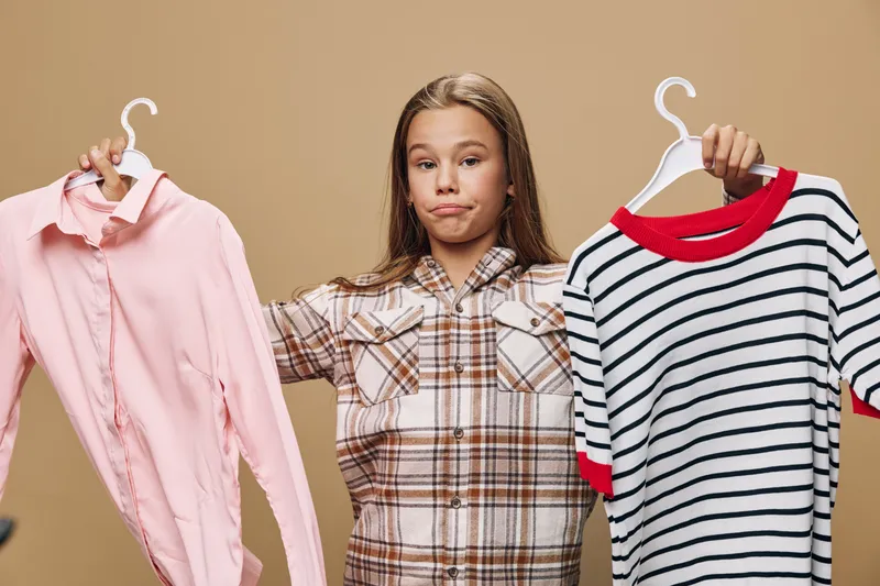 Girl holding two clothes on hangers with indecisive expression comparing pink shirt and striped sweater against beige background in studio.