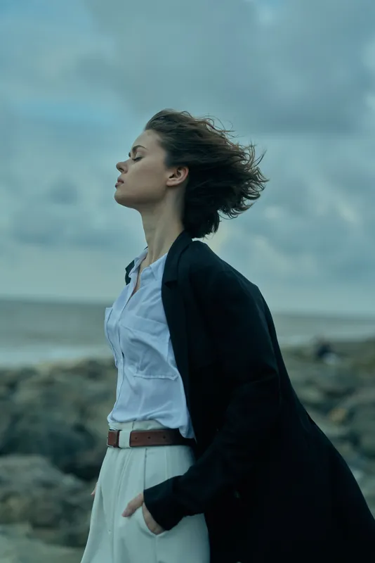 Woman in a flowing coat stands by the ocean, wind swept hair and a calm blue outfit, sea spray and moody clouds create a cinematic outdoor fashion scene