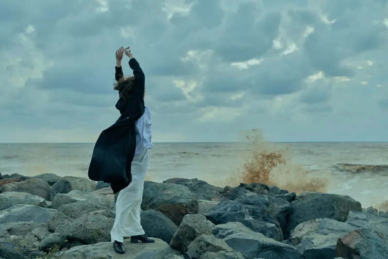 Figure in flowing dress and coat on a rocky shore, arms raised, wind lifting fabric, dramatic motion beside the sea and cloudy horizon, outdoor coastal scene with spray and movement