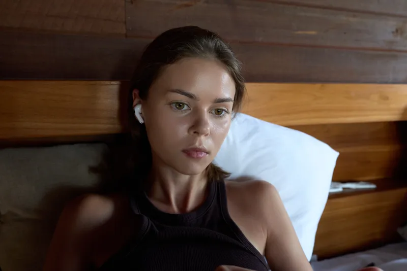 Young woman lying on a bed wearing a black tank top and wireless earphones, looking relaxed with a calm, thoughtful expression as she rests in a quiet indoor setting.