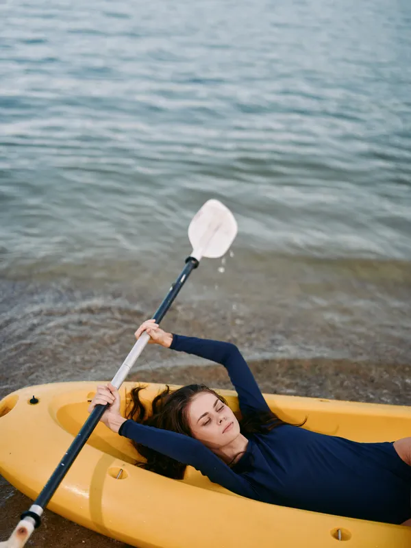 Person reclining in a yellow kayak near a calm shoreline, holding a paddle overhead, sunlit water behind, peaceful moment of outdoor adventure, relaxation, and personal reflection on a warm day.
