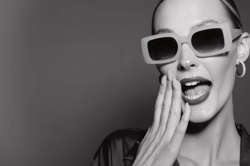 Stylish young white woman with fair skin wearing large sunglasses and hoop earrings covers mouth with hand in black and white studio portrait. Concept of fashion and glamour.