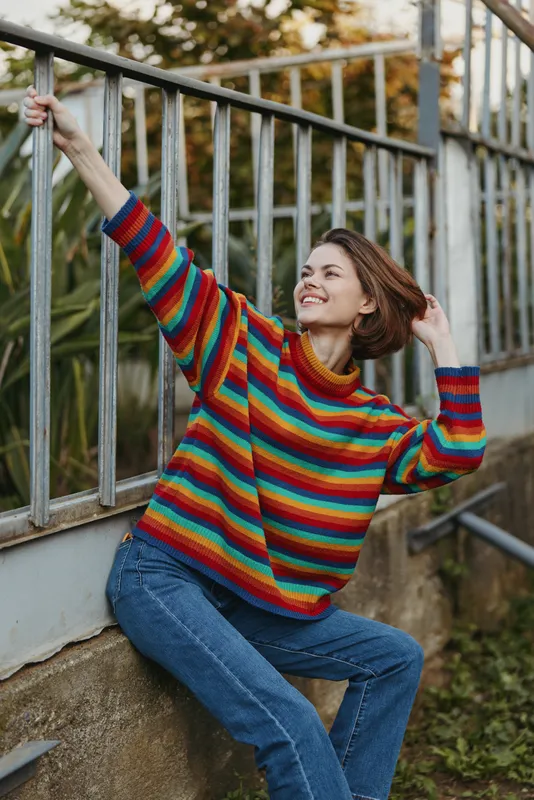Woman in a colorful striped sweater smiling while sitting by metal railing outdoors, casual pose with short hair and blue jeans, relaxed urban portrait in natural autumn light.