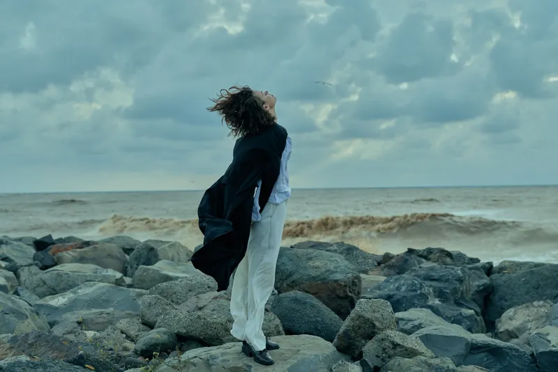 Windy seashore scene with a cloaked figure standing on rocks, hair blown, coat and dress trailing as waves crash and clouds swirl above.
