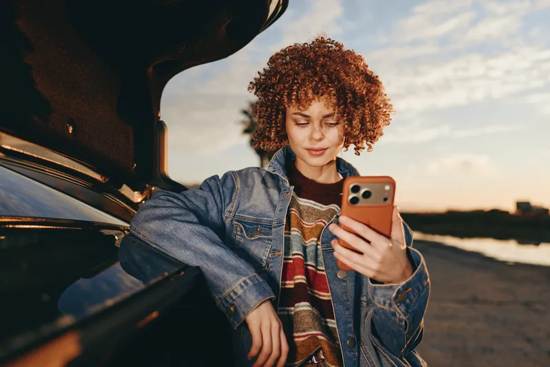 Woman with curly hair taking a selfie with a smartphone near a car, wearing a denim jacket, at sunset. Outdoor lifestyle travel, warm golden light, relaxed smile, evening casual portrait.